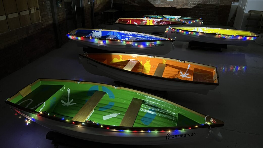 Nine colourful rowing boats displayed indoors, each decorated with painted imagery and strung with multicoloured fairy lights. Boats feature green, orange, blue, red and yellow designs relating to audio stories.