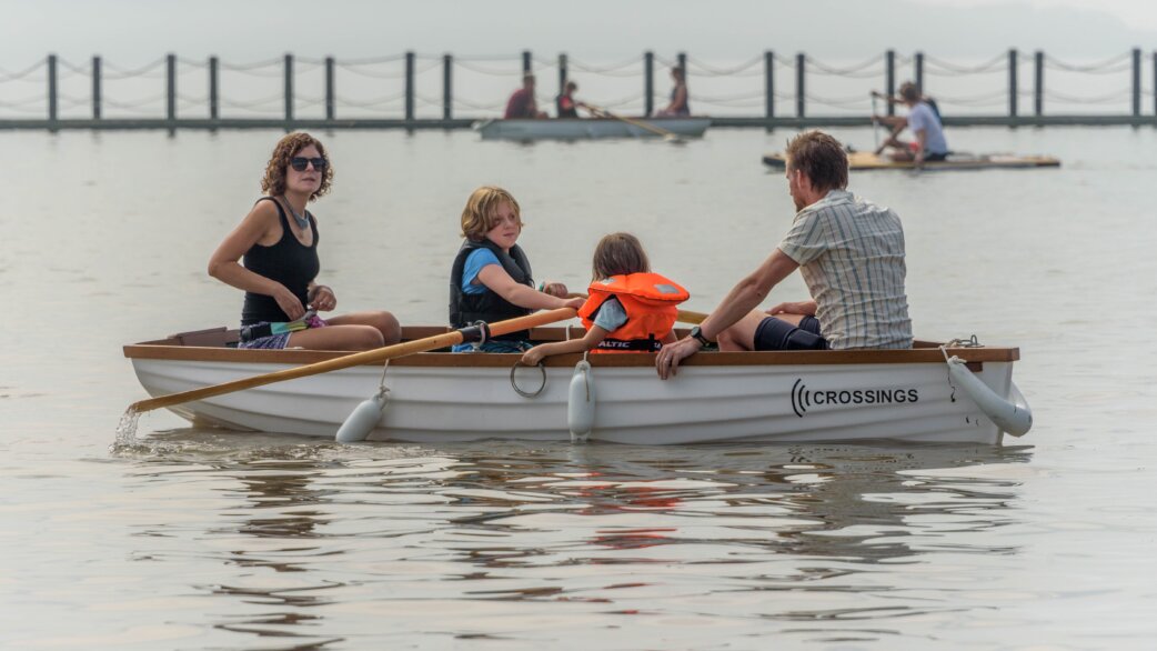 A family of four rows a white wooden boat marked "Crossings" on calm water, with children wearing life jackets and other watercraft visible in the background.
