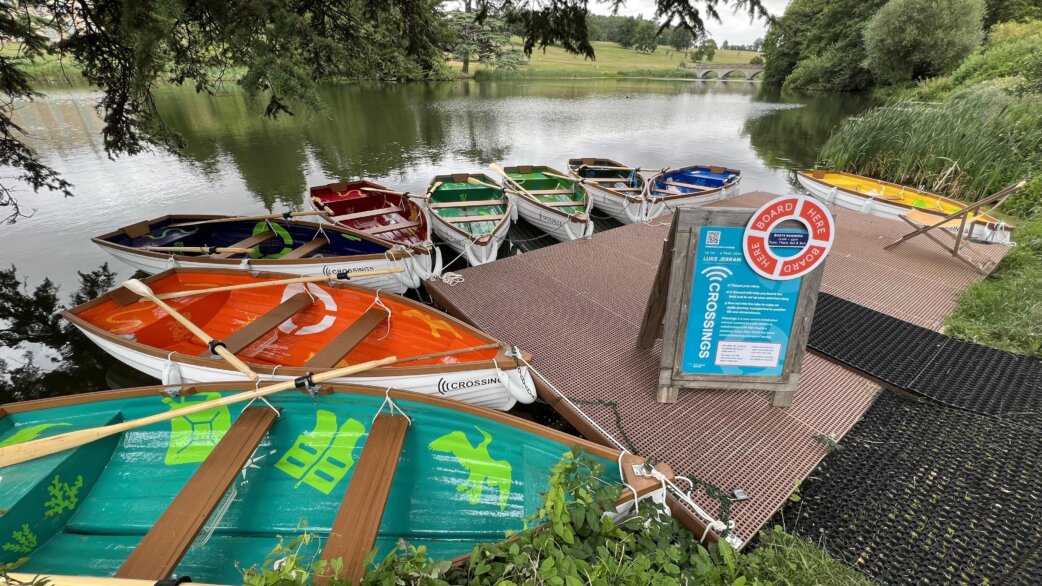Colourful rowing boats moored at a wooden dock on a lake, each decorated with painted imagery. A "Board Here" sign and information board stand on the platform. Trees frame the scene.