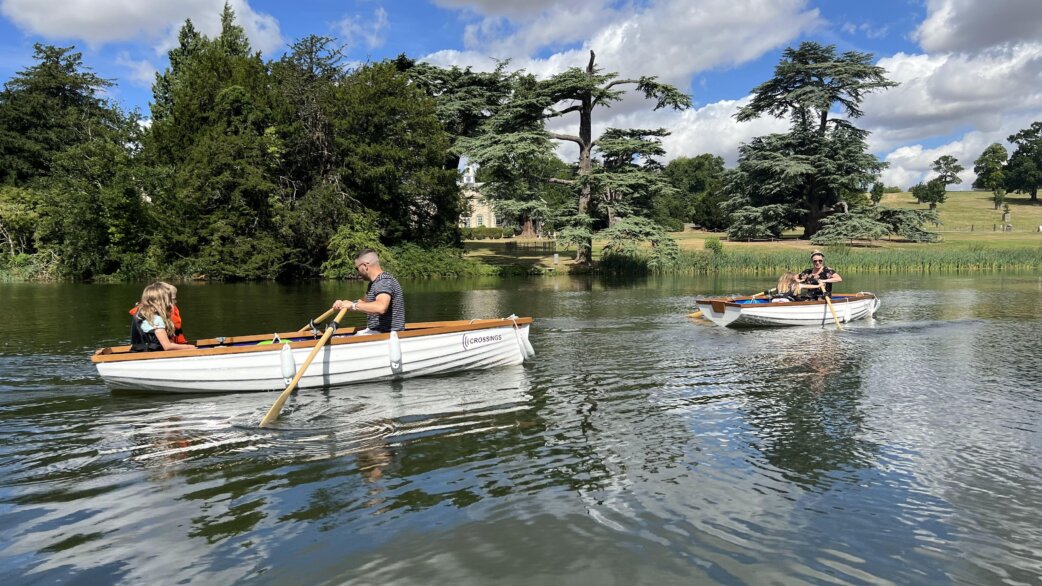 Two white rowing boats on a calm lake, each with passengers rowing while listening to audio stories. Lush green parkland with mature cedar trees and a manor house visible in the background.