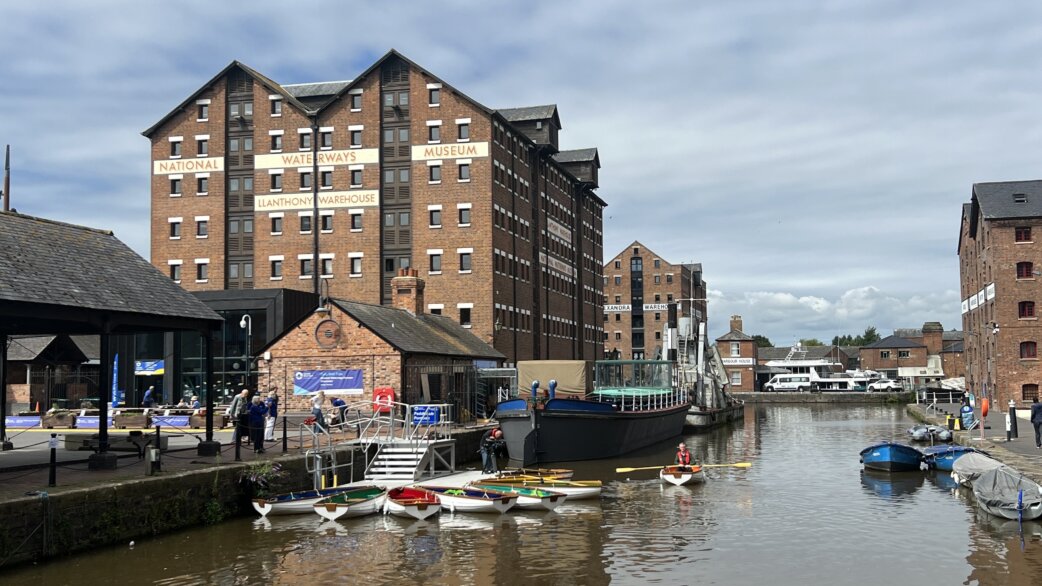 Rowing boats moored in a dock at the National Waterways Museum, Gloucester, with Victorian brick warehouses in the background. The boats are part of Luke Jerram's Crossings installation.