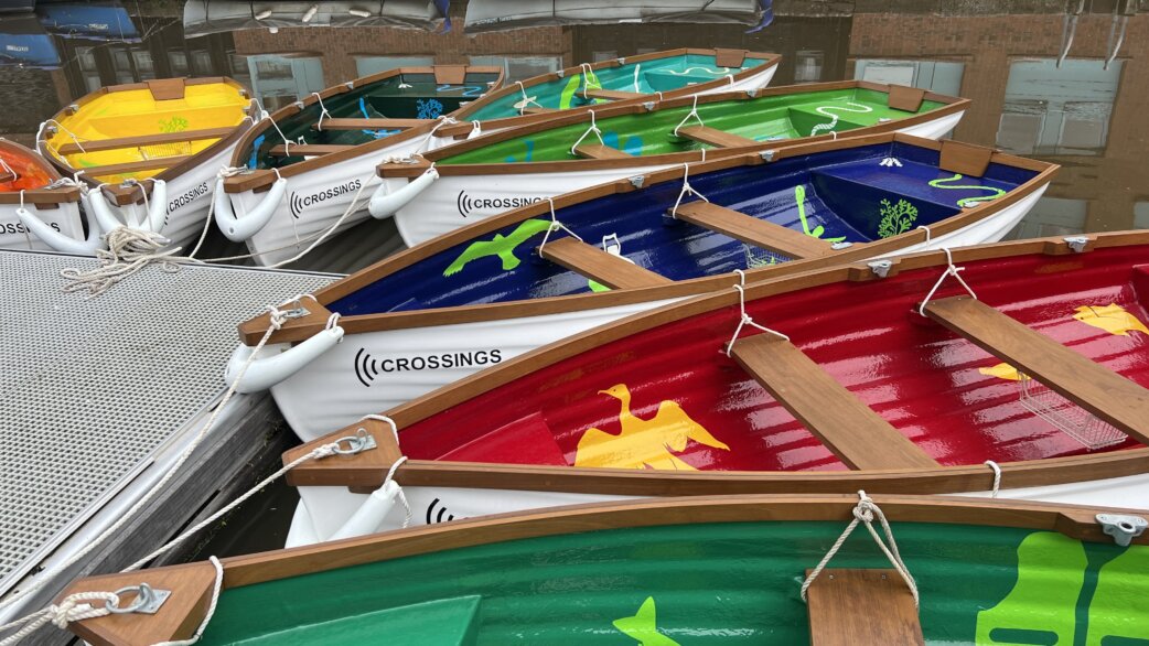 Colourful rowing boats moored together on water, each painted in different colours including red, blue, green and yellow, decorated with animal motifs and branded with "Crossings" logos.