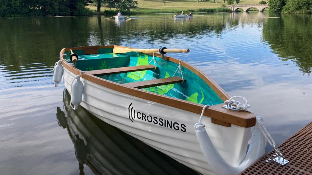 A white rowing boat named "Crossings" moored at a wooden jetty on a calm lake, with wooden oars and a colourful turquoise and green painted interior. Other boats visible in the background.