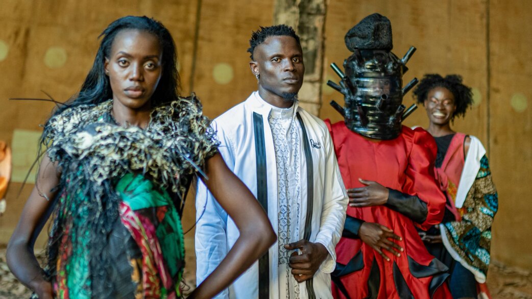 Four models display avant-garde fashion designs in Uganda, featuring elaborate textures, bold colours and an striking sculptural helmet-like headpiece, promoting vaccination awareness through clothing.