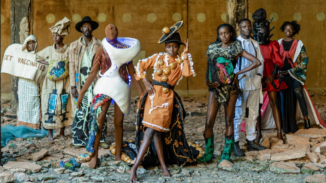 A group of models wearing avant-garde fashion designs made from recycled fabrics, standing in a ruined building. One outfit reads "GET VACCINATED". The clothing features bold colours, sculptural elements and African textiles.