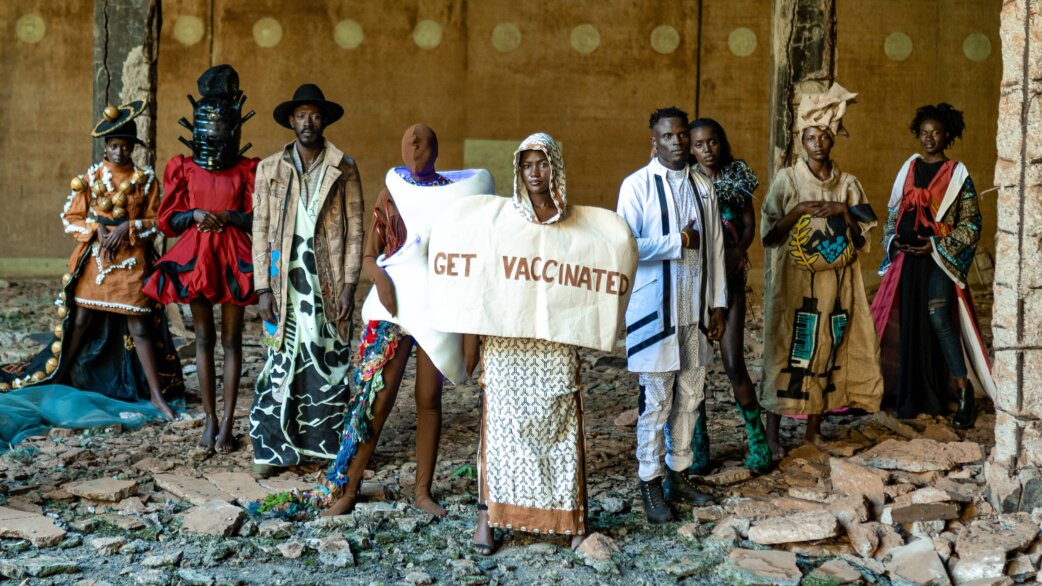 A group of nine people in elaborate recycled fabric garments stand in a ruined building, with a central figure holding a banner reading "GET VACCINATED".