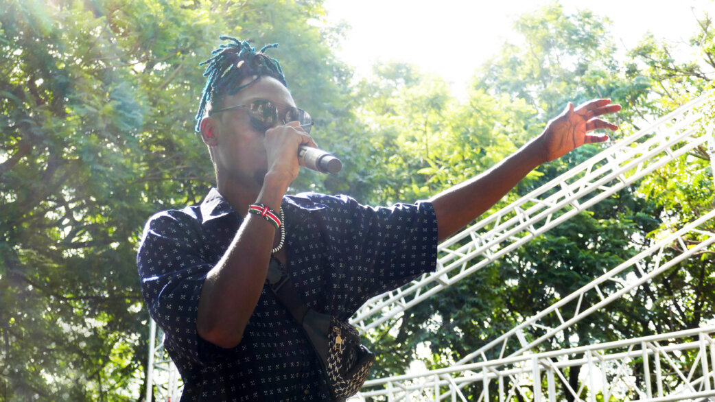 A performer with blue braided hair and sunglasses singing into a microphone at an outdoor event, wearing a dark patterned shirt, with stage scaffolding and trees behind.