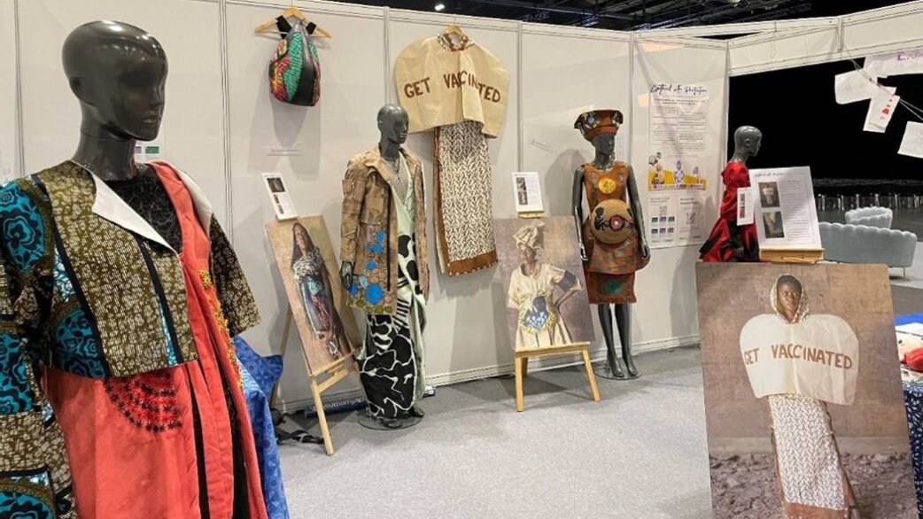 Exhibition display featuring mannequins wearing colourful African-inspired garments made from recycled fabrics, alongside paintings of women in clothing bearing "Get Vaccinated" messaging.