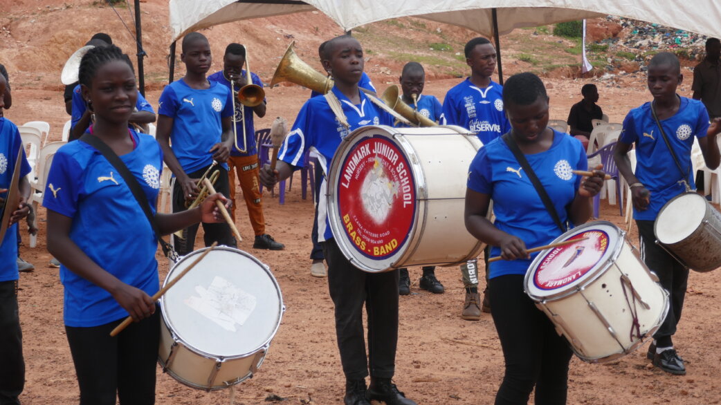 A school brass band from Landmark Junior School performs outdoors on red earth, with young musicians in blue shirts playing drums, trumpets and horns under a canopy.