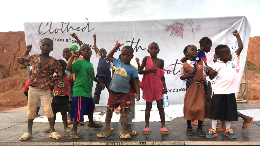 A group of children stand on an outdoor stage in Uganda, raising their fists in celebration, with a banner reading "Clothed with Protection" promoting vaccination behind them.