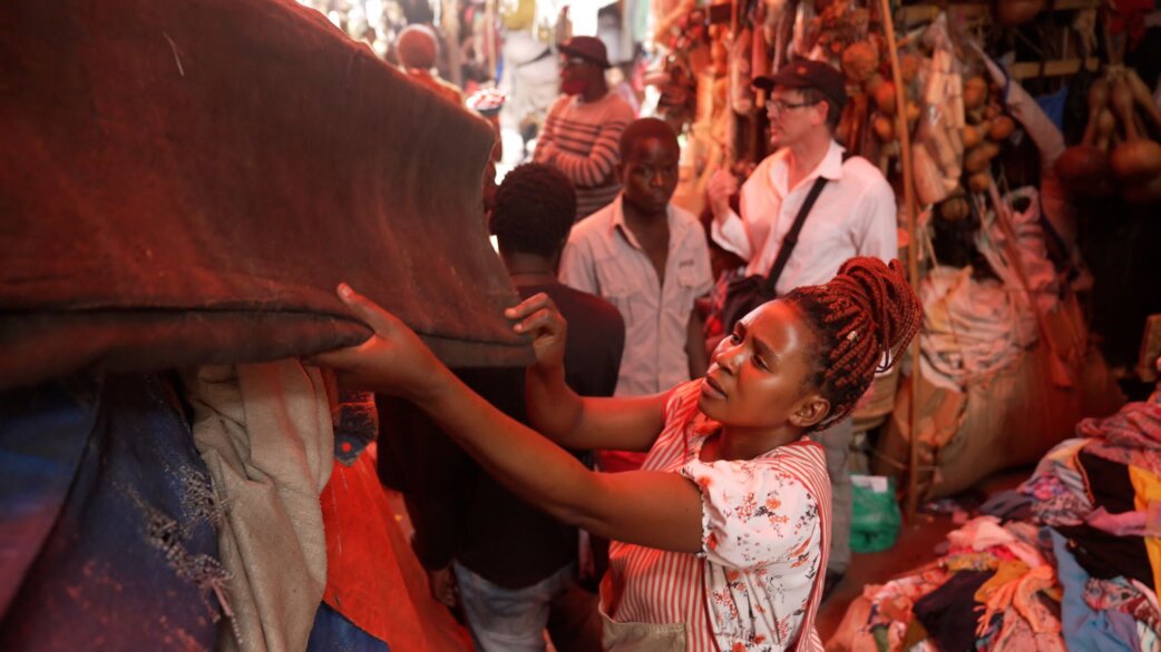 A woman with red braids examines fabric in a busy Ugandan market, surrounded by piles of colourful recycled textiles and other shoppers browsing stalls.