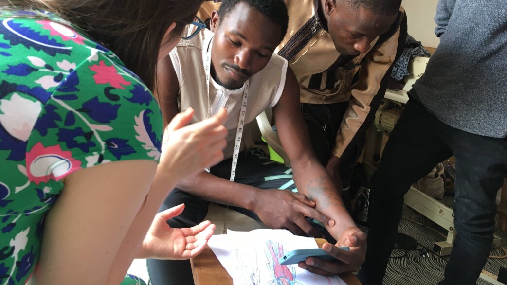 Fashion designers in Kampala, Uganda, collaborate around sketches and fabric samples, with one wearing a measuring tape around his neck, during the Clothed with Protection vaccination awareness project.