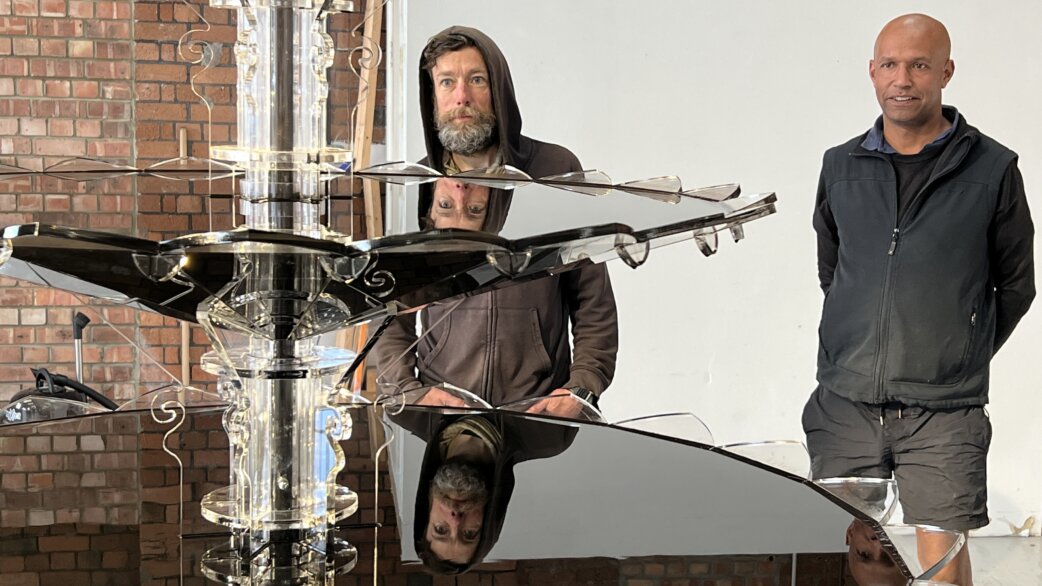 Two men standing beside Oil Fountain, a sculptural piece with a central acrylic column and horizontal mirrored pools reflecting their faces, approximately 1.5 metres tall.