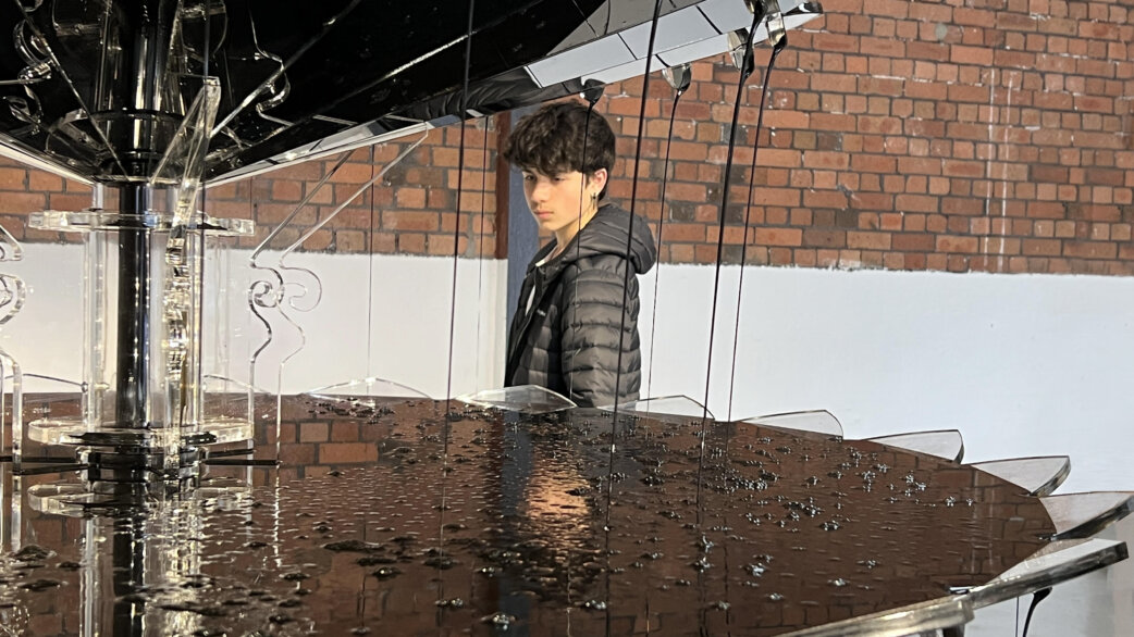 A young person in a dark puffer jacket views Oil Fountain, a sculptural artwork featuring dark engine oil flowing over tiered clear acrylic discs, approximately 1.5 metres tall, against a brick wall.