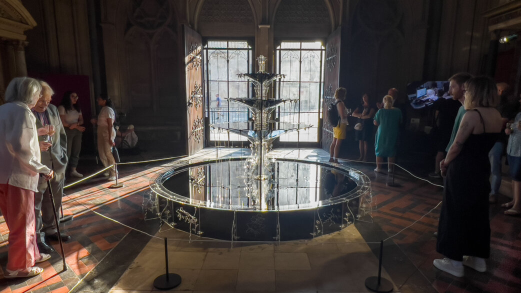 A circular fountain sculpture approximately 2 metres in diameter flows with black engine oil in Bristol Cathedral, surrounded by visitors. Laser-cut acrylic details depict extinct plants and animals.