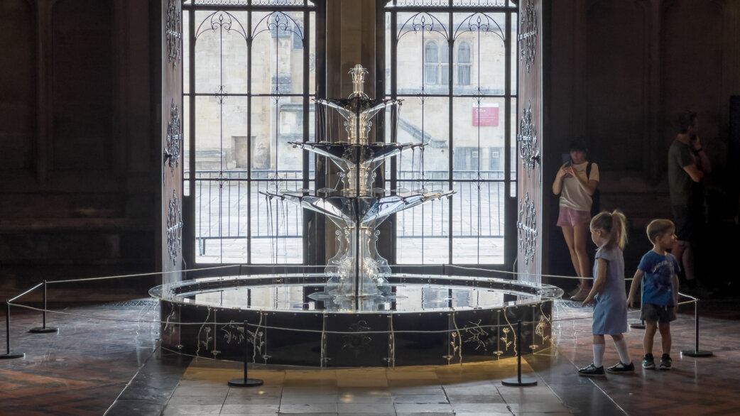 A three-tiered fountain approximately 2 metres tall flowing with dark engine oil, set in Bristol Cathedral with ornate windows behind. Two young children observe nearby.
