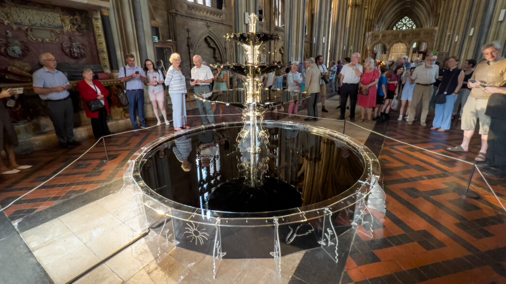 A circular fountain sculpture approximately 2 metres wide, made from clear acrylic filled with dark engine oil, displayed in Bristol Cathedral with visitors gathered around observing.