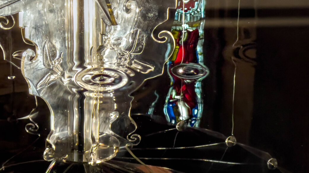 A close-up of a sculptural fountain made from recycled acrylic, with dark engine oil flowing over ornate transparent forms, reflecting colourful stained glass windows from Bristol Cathedral.