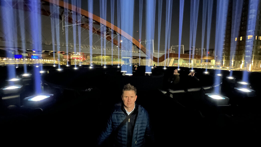 A man stands among an outdoor light installation at night, with numerous ground-mounted spotlights projecting vertical blue-white beams skyward, an arched bridge visible behind.