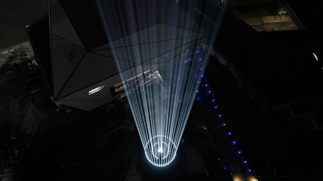 A dramatic installation view showing multiple white light beams radiating upward from a central point in a dark industrial space, with blue LED lights visible along a diagonal line.