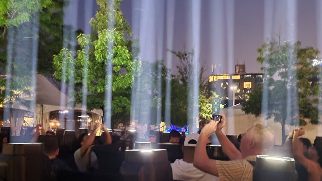 Crowds photograph vertical beams of light shooting skyward at dusk, with green trees and urban buildings visible in the background. The installation creates an ethereal atmosphere.