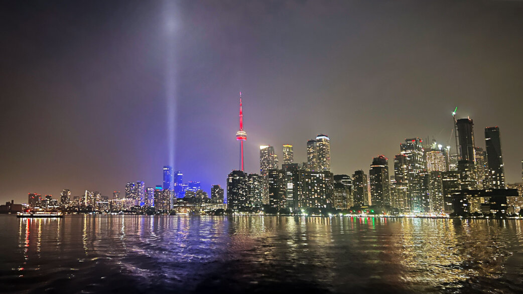 Night view of Toronto skyline from the water, featuring two bright vertical light beams rising into the sky beside the illuminated CN Tower, with city lights reflected on the harbour.