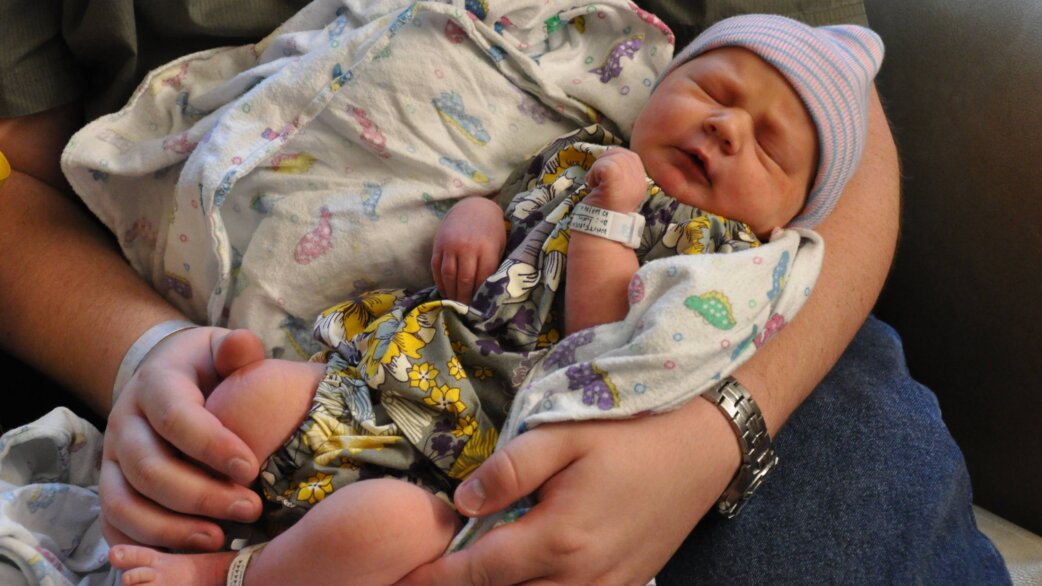 A newborn baby wearing a pink and blue striped hospital cap sleeps peacefully while being cradled in an adult's arms, wrapped in floral-patterned hospital blankets.