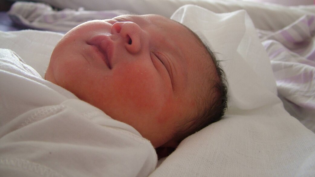 A newborn baby with dark hair and reddish skin lies swaddled in white fabric, eyes closed, appearing to be sleeping peacefully shortly after birth.