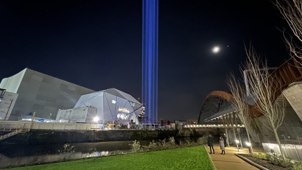 A tall beam of blue light rises vertically into the night sky from behind a modern angular building, with the moon visible and two people walking along a riverside path.
