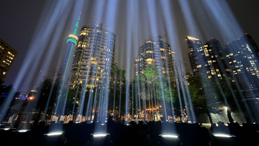 Nighttime light installation featuring multiple vertical white light beams projecting skyward from ground-level spotlights, with Toronto's CN Tower and high-rise buildings visible in the background.