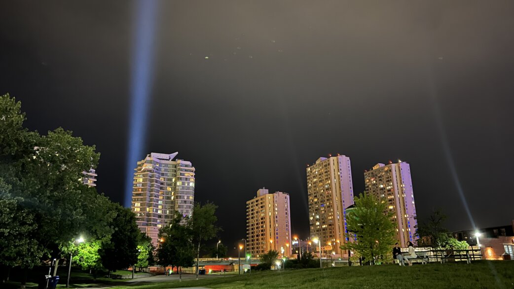 Two vertical beams of light project skyward into a dark night sky above residential tower blocks, viewed from a park with illuminated trees in the foreground.