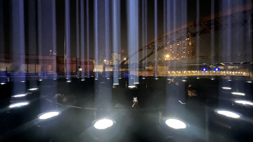 Nighttime light installation featuring multiple upward-pointing searchlights creating vertical beams rising into the dark sky, with visitors walking among the ground-mounted fixtures near an industrial bridge setting.