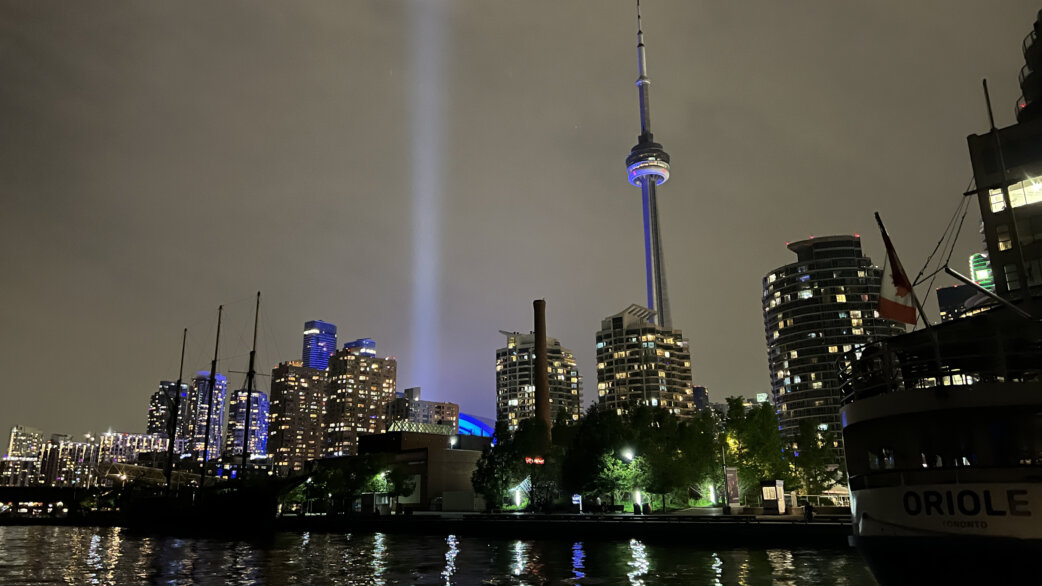 Nighttime Toronto skyline with the CN Tower illuminated, featuring a bright vertical beam of blue light rising into the cloudy sky from the harbourfront area.