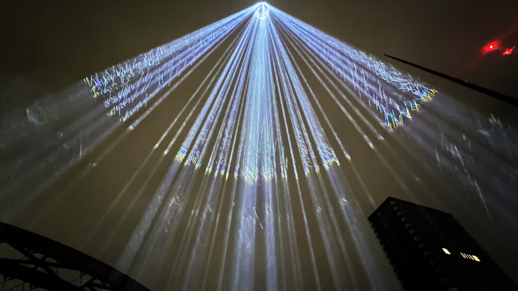 Beams of white light radiate upward into the night sky from a central point, creating a dramatic fan pattern with scattered light effects, viewed from below between dark buildings.