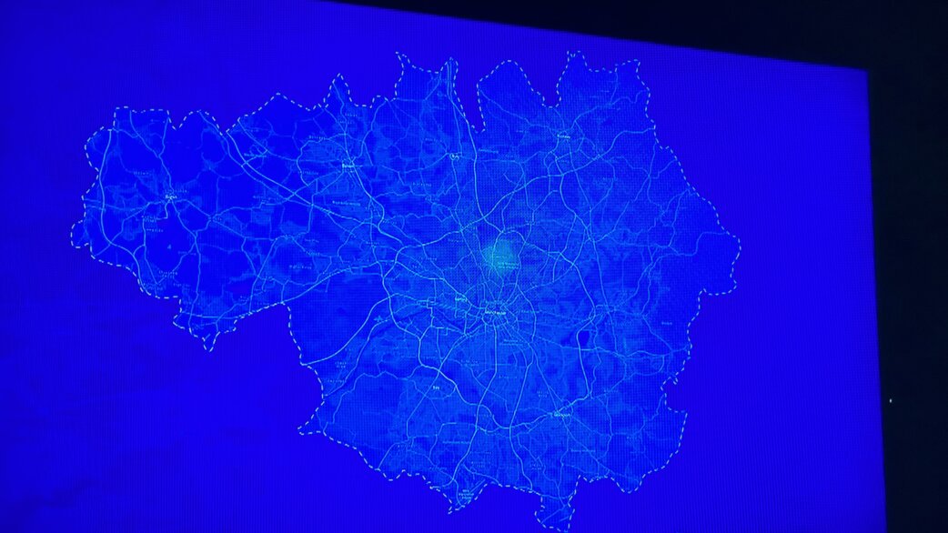 A blue-toned digital map projection showing Greater Manchester's road network and boundaries, with a glowing central point near Manchester city centre, displayed on a screen.