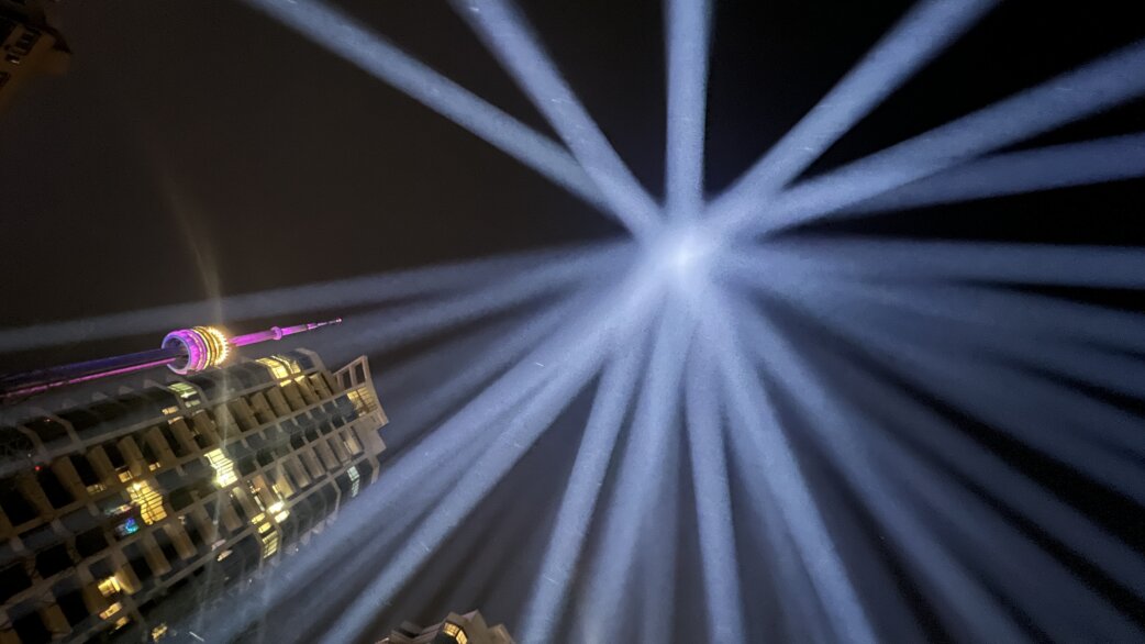 Nighttime photograph of white light beams radiating outward from a central point above a building, with an illuminated tower visible in the background.