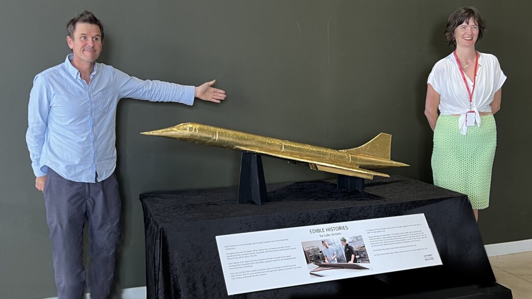 Two people stand beside a gold foil-wrapped chocolate sculpture of Concorde, approximately 1.5 metres long, displayed on a black-clothed table with an information panel reading "Edible Histories by Luke Jerram".