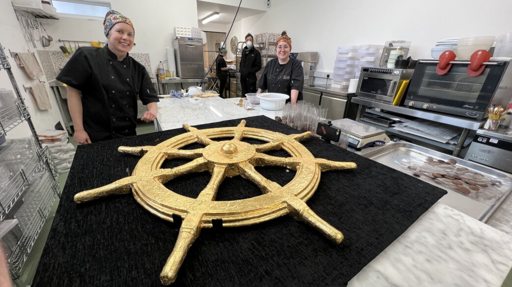 A gold foil-wrapped chocolate ship's wheel, approximately one metre in diameter, displayed on a black surface in a commercial kitchen, with three smiling staff members nearby.