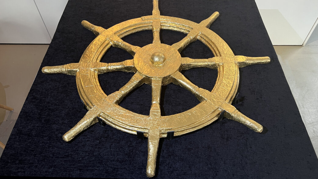 A golden foil-wrapped chocolate replica of a ship's wheel, approximately one metre in diameter, displayed on dark blue velvet fabric. The wheel has eight spokes and textured surface detail.