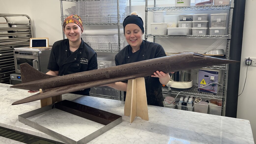 Two chocolatiers in black uniforms smile while displaying a chocolate replica of Concorde, approximately 1 metre long, mounted on wooden stands in a professional kitchen.