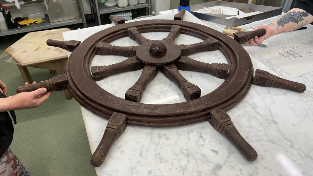 A chocolate ship's wheel, approximately one metre in diameter, held by two people in a workshop setting. The wheel has eight spokes and shows realistic weathered detailing.