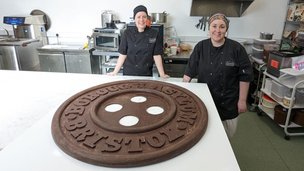 Two chocolatiers in black uniforms stand in a commercial kitchen behind a 1-metre diameter chocolate replica of a Bristol Borough Asylum button with four holes.