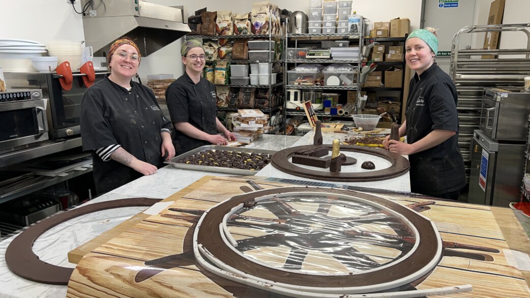 Three chocolatiers in black uniforms stand in a commercial kitchen with large chocolate ship's wheels, approximately one metre in diameter, displayed on wooden boards.