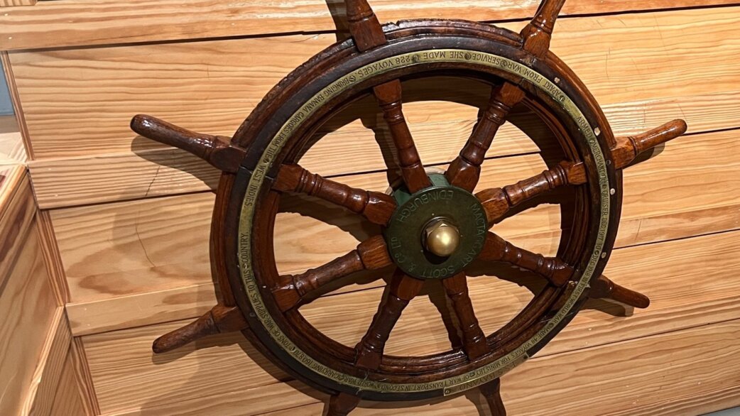 A wooden ship's wheel approximately one metre in diameter with eight spokes, brass centre hub and inscribed brass ring, displayed against wooden panelling.