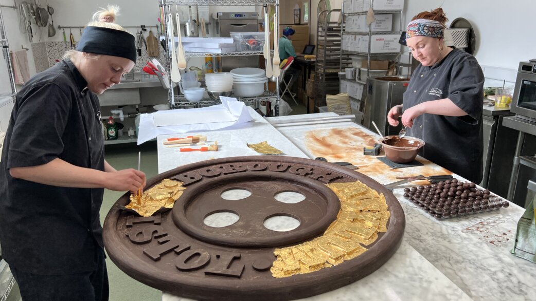 Two chocolatiers in a kitchen applying gold foil to a large chocolate replica of a Victorian asylum button, approximately one metre in diameter, with raised text reading "Borough Asylum".