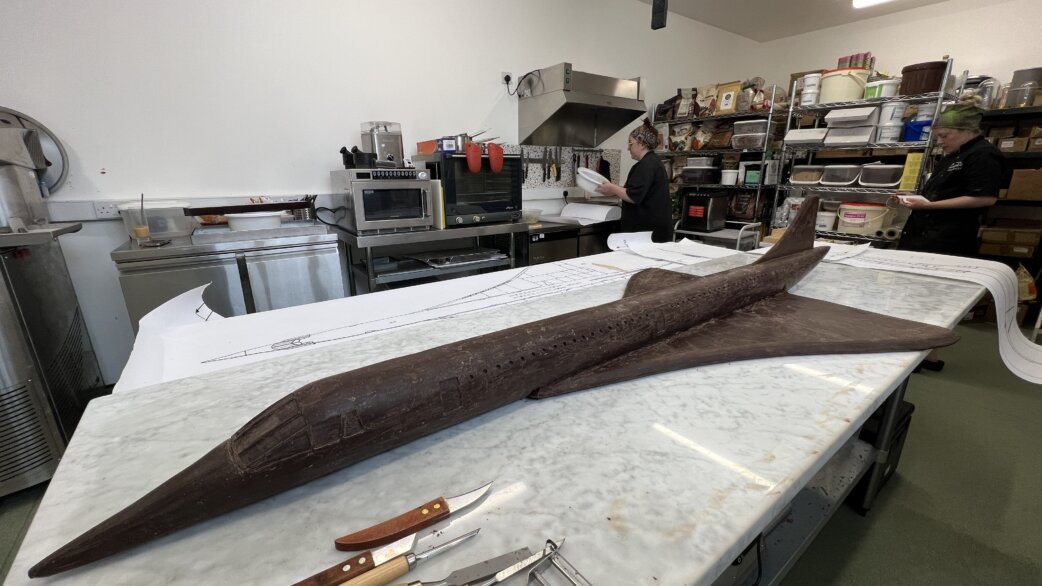 A chocolate sculpture of Concorde, approximately 1.5 metres long, resting on a marble worktop in a commercial kitchen, with blueprints visible beneath and staff working in the background.