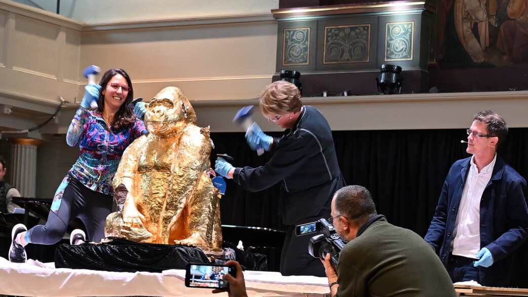 Two people wearing blue gloves use hammers to break apart a gold foil-wrapped chocolate gorilla sculpture, approximately one metre tall, on a stage while onlookers photograph the event.