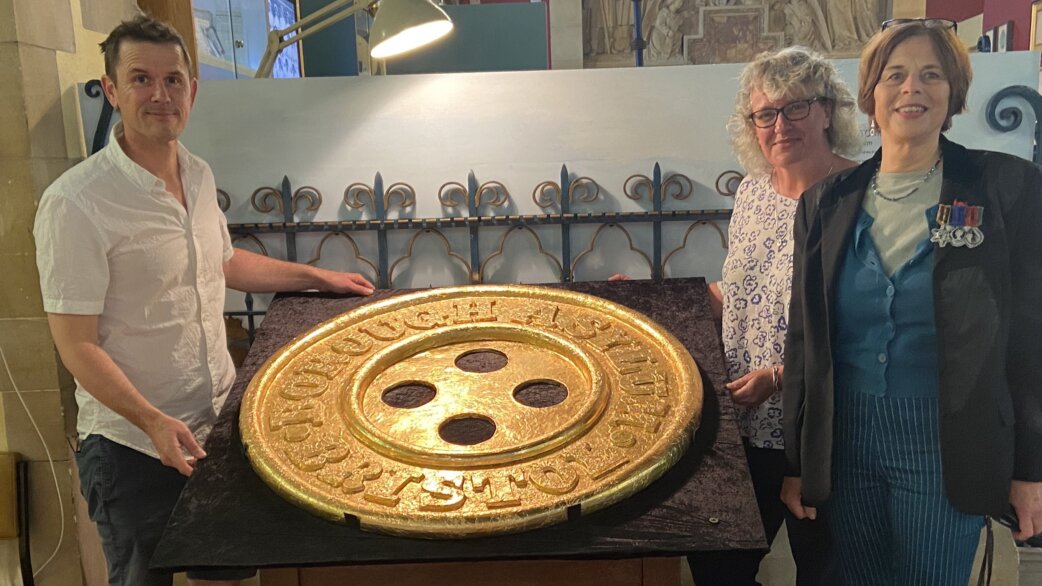 Three people stand beside a large gold foil-wrapped chocolate button sculpture, approximately one metre in diameter, featuring embossed text around its rim and four central holes.