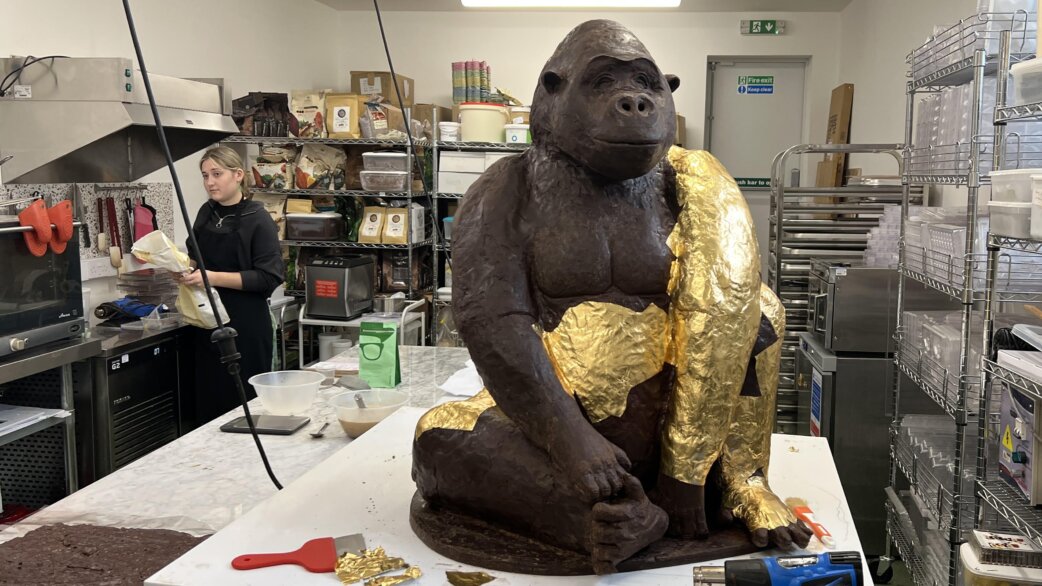 A life-sized chocolate gorilla sculpture with gold foil detailing on its arms and chest, positioned on a worktop in a commercial kitchen, with a chocolatier working in the background.