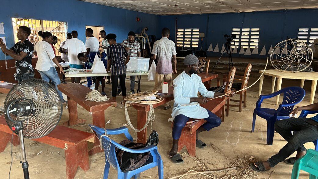 Workshop in Freetown, Sierra Leone showing participants constructing paper lantern frames from cane and wire, with completed spherical structures visible in background. Blue-walled community space.